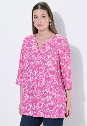 Kobieta o długich prostych włosach, nosząca perłowe kolczyki i różową tunikę w kwiatowy wzór z rękawami do łokcia.