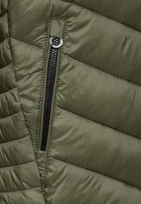 Veste rembourrée verte avec des sections matelassées horizontales, une fermeture éclair noire avec un tirette circulaire et une texture de tissu lisse et brillante.