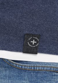 T-shirt en tissu côtelé bleu marine avec une étiquette noire portant l'inscription "Key Largo" cousue sur le col, superposée sur une chemise blanche et un jean bleu.