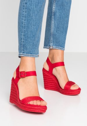Czerwone sandały na koturnie z plecioną fakturą, z dwoma szerokimi paskami i regulowanym paskiem wokół kostki, na tle dżinsów.