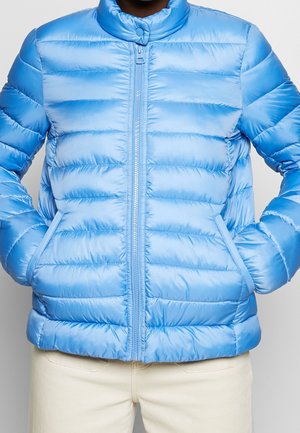 Persona que lleva una chaqueta acolchada azul claro con cremallera frontal y bolsillos, combinada con pantalones color marfil.