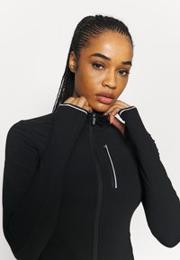 Musta urheilutakki, jossa on korkea kaulus, vetoketjullinen etutasku ja sormeen laitettavat aukot hihoissa. Pehmeä, joustava kangas, jossa on hienovaraisia valkoisia aksentteja.