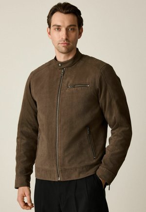Man wearing brown suede zip-up jacket with chest and side zip pockets, standing against plain light background.