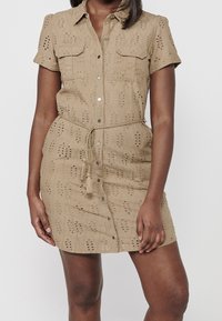 Vestito beige a maniche corte con dettagli ricamati e due tasche frontali. Presenta una chiusura frontale con bottoni e una vita con cintura e frange.