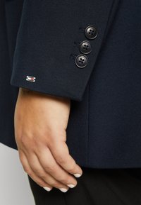 Poignet de blazer marine avec trois boutons noirs portant l'inscription "TOMMY" en relief. Comprend une petite étiquette de marque en rouge et blanc sur la manche.