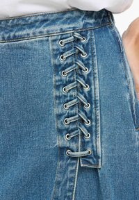 Jeansrock in Hellblau mit einer Reihe silberner Ösen und Schnürung an der Seite. Weiche Textur und markante Nahtdetails.