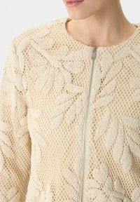 Beżowa kurtka o fakturze z kwiatowym haftem, z okrągłym dekoltem i zapięciem na zamek. Tkanina ma siatkowy wzór.