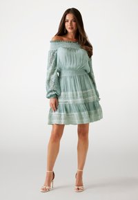Mintgrünes Off-Shoulder-Kleid mit langen Spitzenärmeln, strukturiertem Stoff, gestuftem Rock und dekorativen Bändern. Kombiniert mit weißen, geschnallten Absätzen.