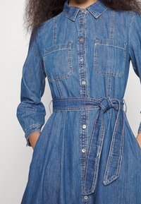 Denim shirt dress featuring a collar, two chest pockets, a waist tie, and button closure; blue fabric with contrasting stitching details.