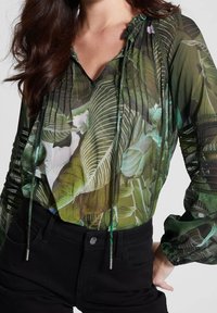 Nainen, jolla on päällään läpikuultava vihreä paita, jossa on trooppinen lehtikuvio, sekä mustat korkeavyötäröiset housut, poseeraa yksi käsi lantiolla.