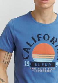 Zila zila kokvilnas T-kreklu ar apļveida grafiku, kurā redzams saulriets oranžos un zilos svītrās, kā arī teksts "California Blend" un "Huntington Beach Longboards".