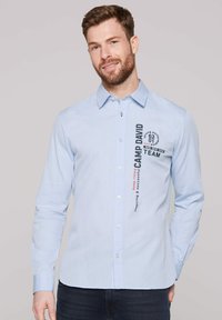 Himmelblaues Langarmhemd mit Knöpfen und subtiler Textur. Verfügt über bedruckte Texte und Grafiken auf der Vorderseite, einschließlich des Markennamens "CAMP DAVID."