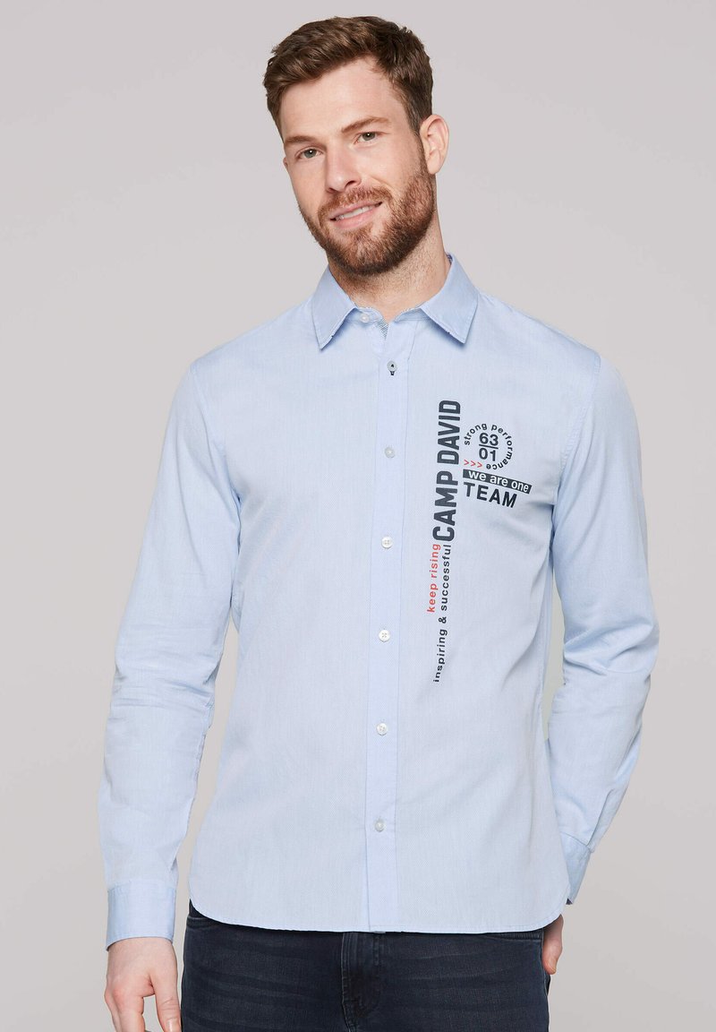 Himmelblaues Langarmhemd mit Knöpfen und subtiler Textur. Verfügt über bedruckte Texte und Grafiken auf der Vorderseite, einschließlich des Markennamens "CAMP DAVID."