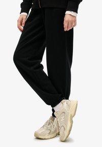 Pantalones de chándal negros de algodón con puños en los tobillos, combinados con zapatillas beige de New Balance que presentan detalles de malla y goma. Se ven calcetines blancos.