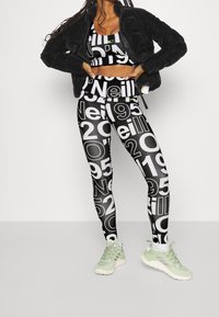 Svart puffjacka, svart och vit mönstrad crop top, och leggings med stora texter. Modellen har på sig ljusgröna sneakers. Slät tyg.