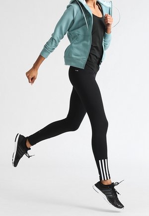Personne en leggings de sport noirs avec des rayures blanches et des baskets noires en train de courir, portant un sweat à capuche zippé turquoise et un débardeur noir.