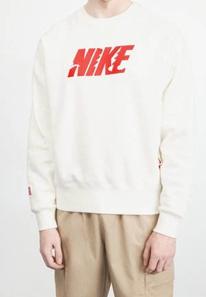 Person trägt einen weißen Sweatshirt mit fettgedrucktem roten "NIKE"-Schriftzug auf der Brust, kombiniert mit beigen Hosen, stehend vor einem schlichten weißen Hintergrund.
