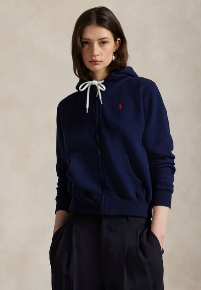 Jeune femme portant un sweat à capuche zippé bleu marine avec des cordons blancs et un pantalon noir, debout les mains dans les poches sur un fond uni.