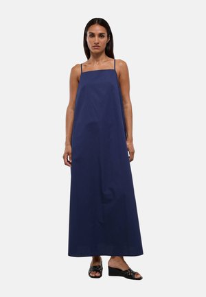 Femme portant une robe bleu marine sans manches, longue jusqu'au sol, avec de fines bretelles et des sandales noires à talons, debout devant un fond uni.