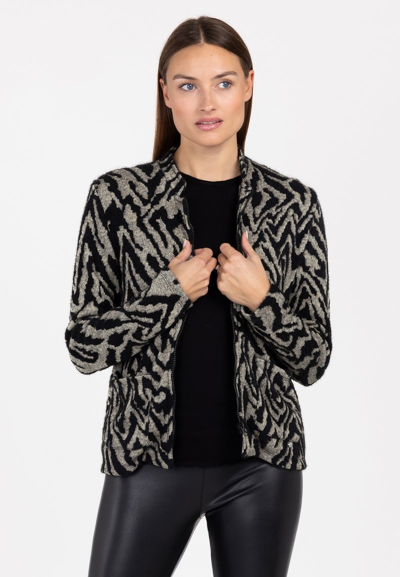 Cardigan a motivi neri e crema con chiusura a zip, polsini a costine e scollatura rotonda, indossato sopra un top nero con un design astratto.