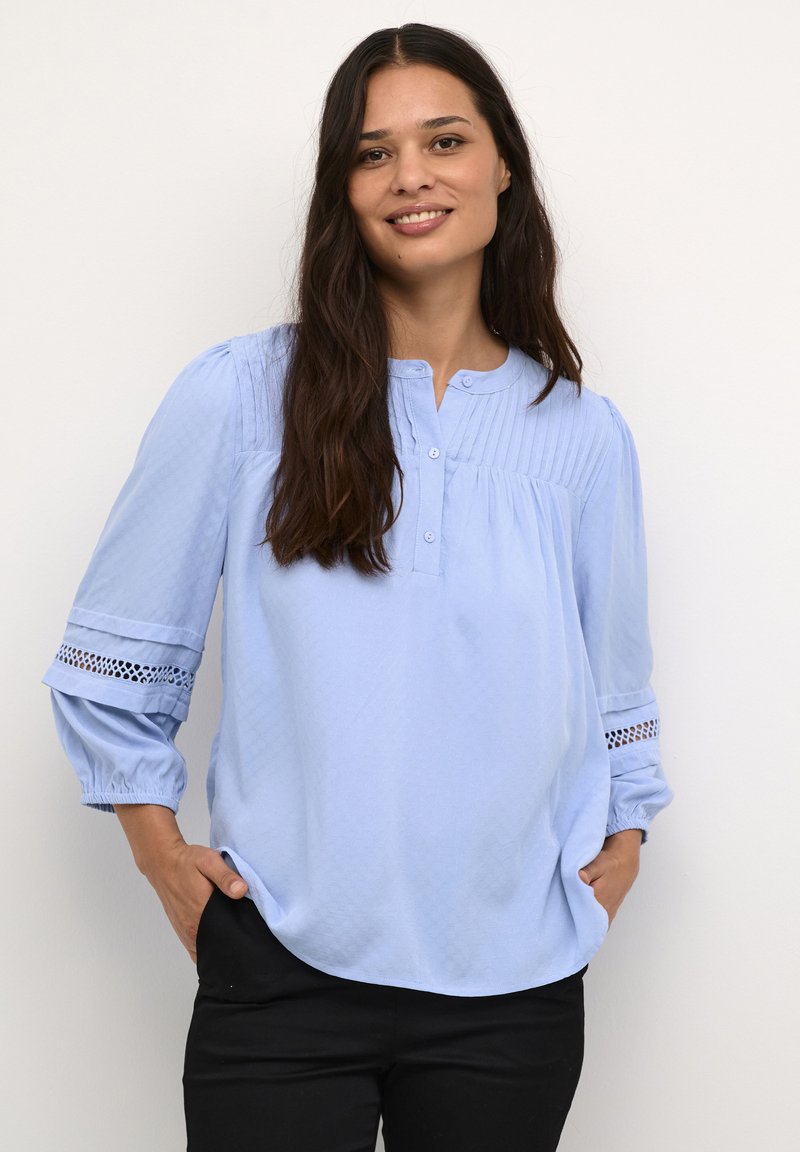 Blusa azzurro chiaro, con texture, dal collo rotondo, chiusura a bottone e maniche a tre quarti con dettagli in pizzo.