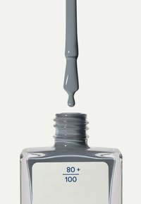 Grå nagellack i en rektangulär glasflaska med ett rundat lock. En droppe lack hänger från borsten ovanför flasköppningen.