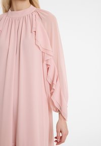 Helles rosafarbenes Kleid aus fließendem Stoff mit gerafftem Ausschnitt und cascadelnden Rüschen an den langen Ärmeln. Weiche Textur und elegantes Design.