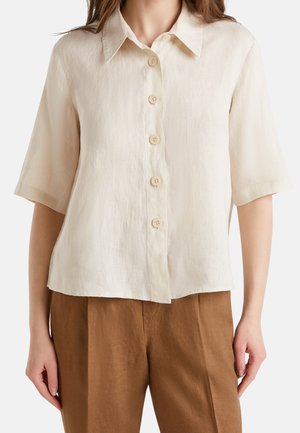 Woman wearing a short-sleeve beige button-up shirt with a collar paired with high-waisted brown pants, against a plain background.