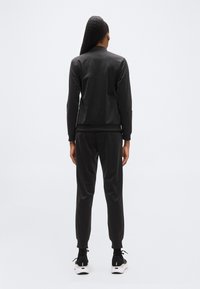 Μαύρο αθλητικό σετ με εφαρμοστό μπουφάν με ριμπ τελείωμα και ταιριαστό παντελόνι με κωνικό κόψιμο. Λείο ύφασμα, συνδυασμένο με μαύρα παπούτσια.