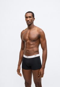 Chiloți boxer negri cu o talie albă pe care este imprimat logo-ul „BOSS”. Materialul pare neted și se potrivește strâns pe corp. Modelul este prezentat fără tricou.