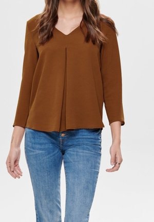 Kobieta ubrana w brązową bluzkę z długim rękawem i dekoltem w serek z plisowanym detalem z przodu oraz niebieskie dżinsy, stojąca na neutralnym tle.