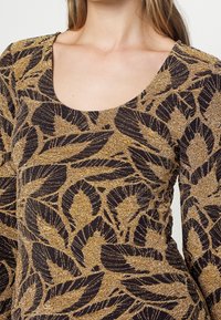 T-shirt à manches longues avec un col bateau, présentant un motif de feuilles noires et dorées sur un tissu texturé pailleté.