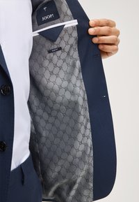 Giacca da abito blu navy con vestibilità slim. L'interno presenta una fodera a motivo grigio e un'etichetta con la scritta "JOOP!". Chiusura frontale con bottone e tessuto testurizzato.