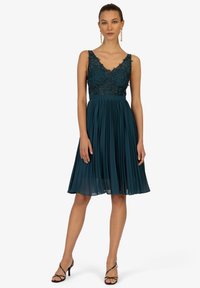 Abito teal con un corpino in pizzo aderente e maniche a cappuccio, caratterizzato da una gonna plissettata che arriva sopra il ginocchio. Tacchi neri con cinturini completano il look.