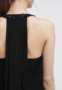 Black sleeveless top with a draped back design, featuring two circular metal accents at the neck and a soft, smooth texture.