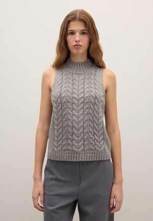 Frau mit welligem Haar trägt einen ärmellosen grauen Zopfpullover mit Rollkragen und graue Hose vor schlichtem hellem Hintergrund.