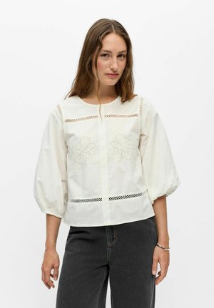 Femme portant un chemisier blanc avec des détails en dentelle florale et des manches bouffantes, associé à un jean gris foncé sur un fond blanc uni.