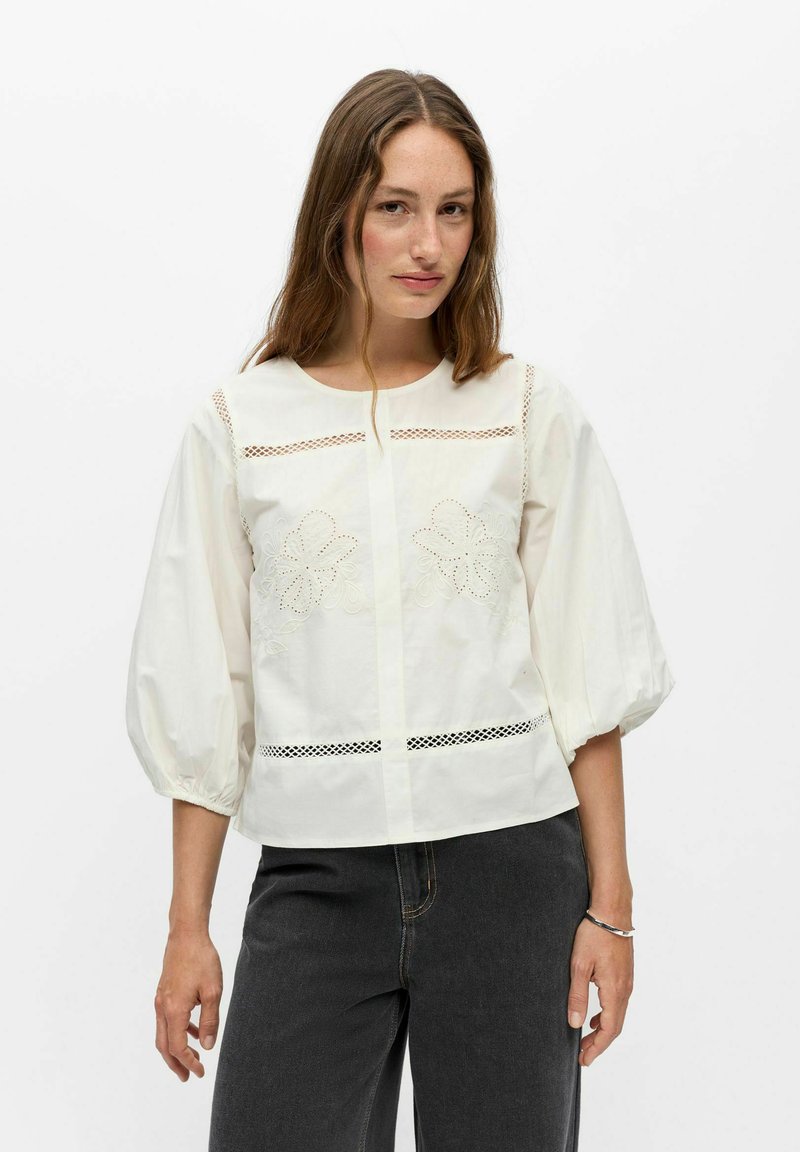 Femme portant un chemisier blanc avec des détails en dentelle florale et des manches bouffantes, associé à un jean gris foncé sur un fond blanc uni.