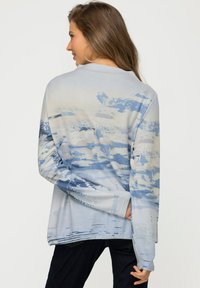 Haut à manches longues bleu clair présentant un design de paysage aquarelle avec de douces vagues et des dégradés de bleus et de crème. Texture lisse.