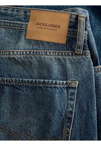 Jeans din denim de culoare albastru mediu, cu o etichetă din piele maro care spune "JACK & JONES", cusături vizibile și un design clasic al buzunarelor.