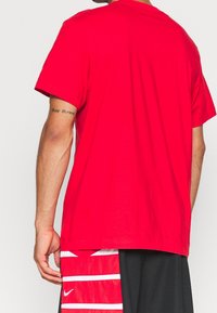 Camiseta de algodón roja con mangas cortas, escote redondo y un diseño liso. Mostrada desde la parte de atrás, combinada con pantalones cortos negros con rayas blancas.