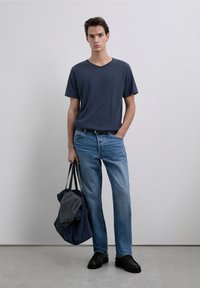 Camiseta de cuello en V azul marino, vaqueros de pierna recta azul claro, cinturón negro, zapatos negros, sosteniendo una bolsa oscura con un suéter gris. Fondo sencillo.