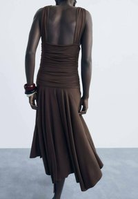 Robe sans manches marron avec un corsage froncé et une jupe évasée. Tissu lisse au design fluide, agrémenté de bracelets de poignet audacieux.
