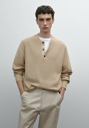 Maglione a coste beige con collo henley, caratterizzato da tre bottoni scuri. Abbinato a pantaloni beige chiaro, creando un outfit neutro.