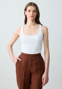Weißes geripptes Tanktop mit quadratischem Ausschnitt, kombiniert mit hochtaillierten braunen Leinenhosen, die Falten vorne und Eingrifftaschen haben.