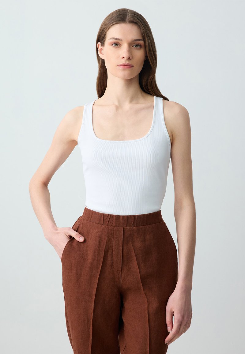 Weißes geripptes Tanktop mit quadratischem Ausschnitt, kombiniert mit hochtaillierten braunen Leinenhosen, die Falten vorne und Eingrifftaschen haben.
