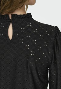 Black blouse featuring a textured fabric with eyelet holes, ruffled neckline, and a keyhole opening at the back. Long puff sleeves.