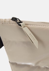 Sac imperméable beige avec une finition texturée, présentant un clip noir et une étiquette marquée "RAINS" attachée par une corde.
