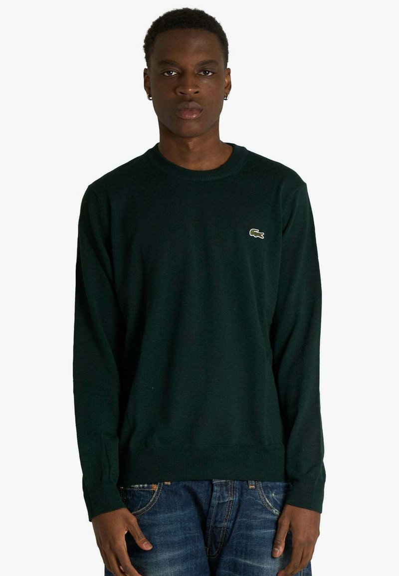 Temno zeleni pulover s krožnim izrezom iz mehke tkanine, z dolgimi rokavi in majhno vezeno logotipom na prsih. Nošen z modrimi jeans hlačami.