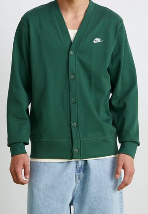 Grøn knap-op cardigan med hvid Nike-logo på venstre bryst, båret over en hvid skjorte, kombineret med lyseblå jeans.